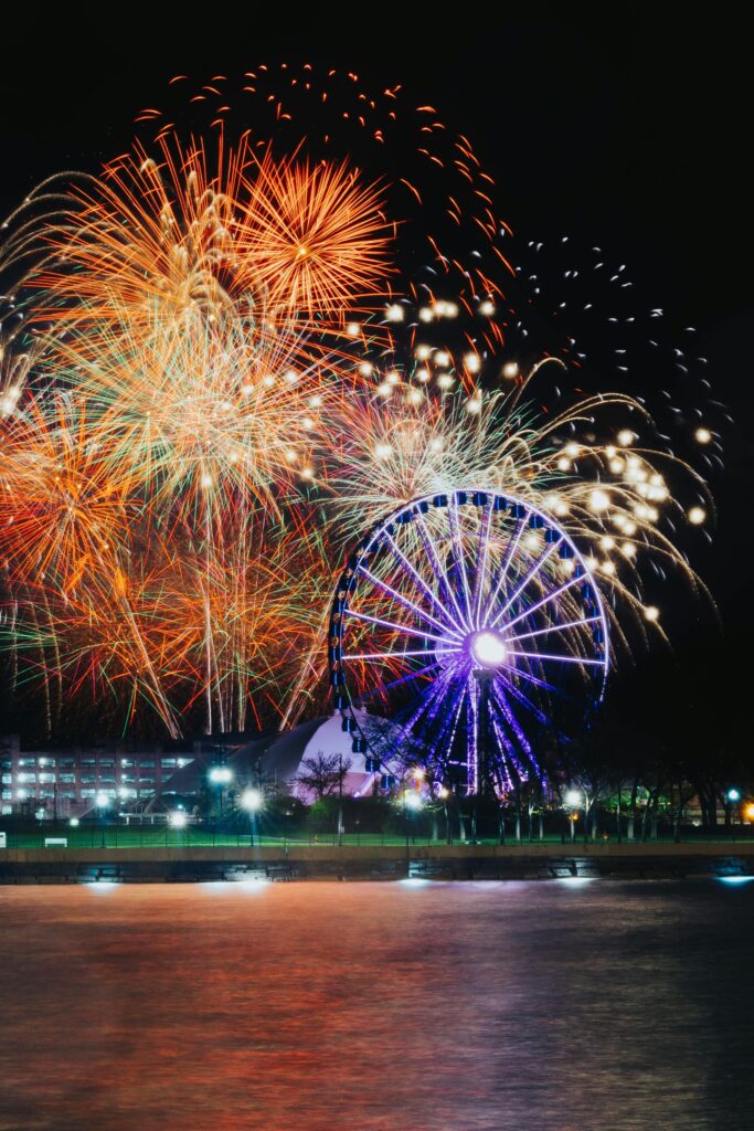 Colorful fireworks illuminate the sky over a Ferris wheel reflection on water during a night celebration.